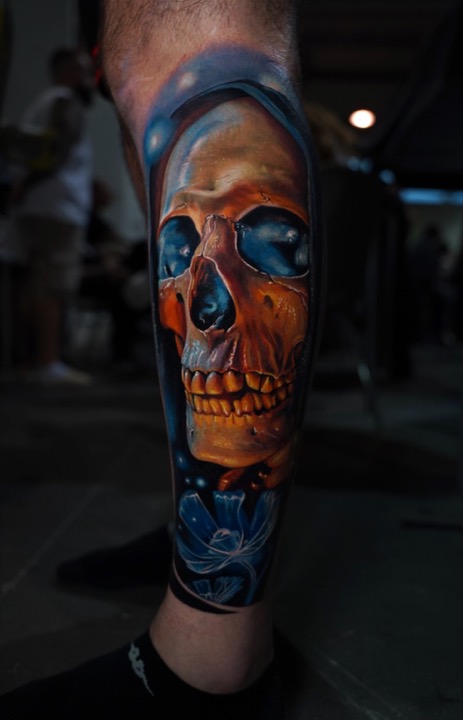 Golden skull with blue eye sockets inside a blue hood, colour tattoo on a calf at a convention.