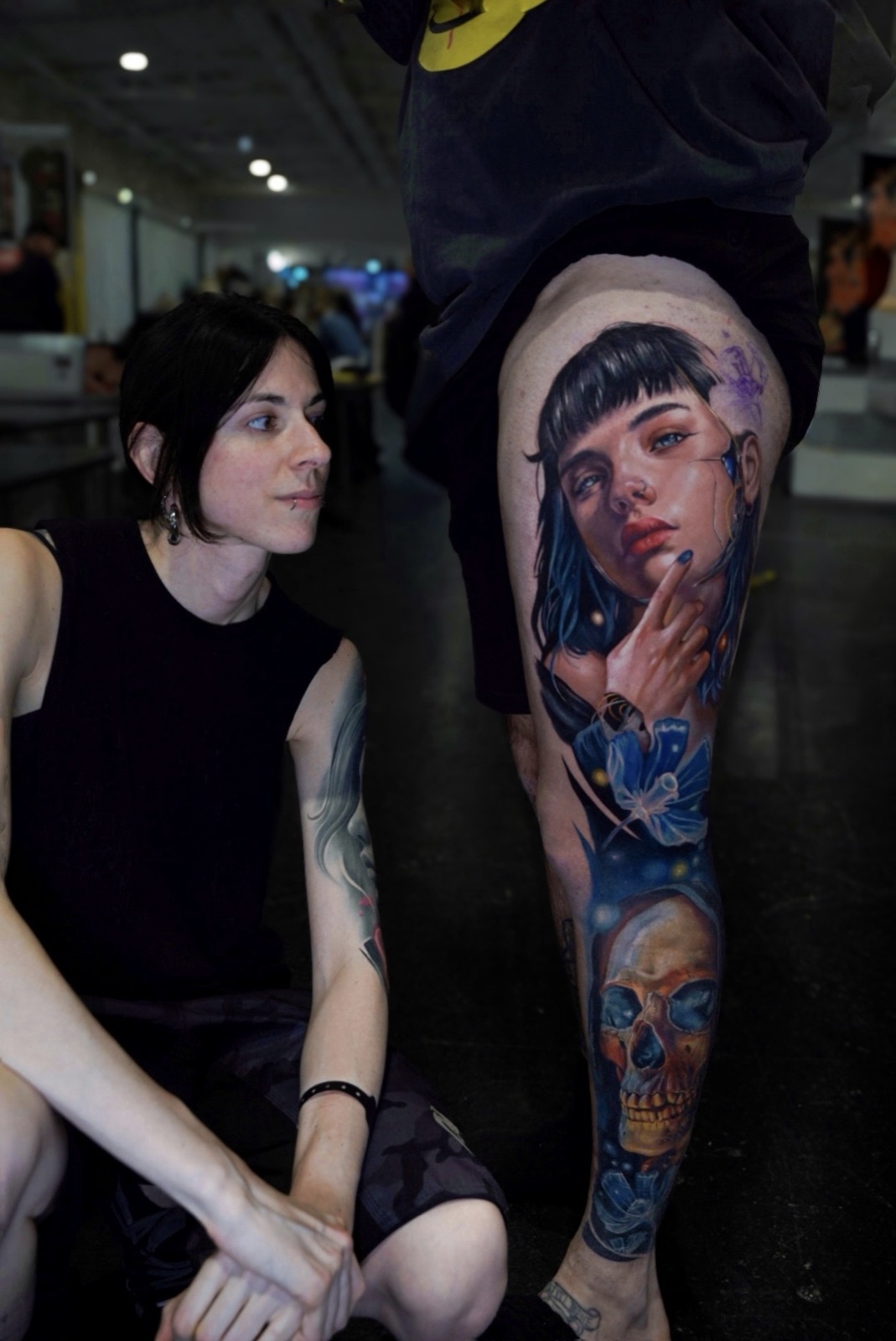 Carlotta crouching beside her client at a tattoo convention, next to a finished colour leg piece: a woman's portrait with a blue butterfly and a golden skull below.