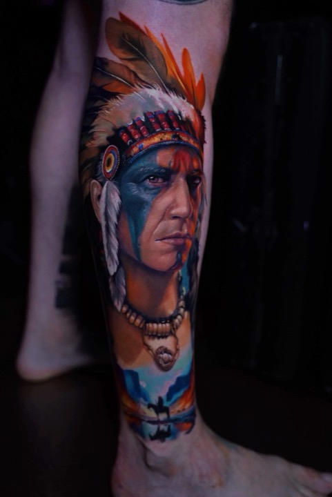Portrait of a man in a feathered headdress with blue face paint and a rider at sunset below, colour tattoo on a calf.
