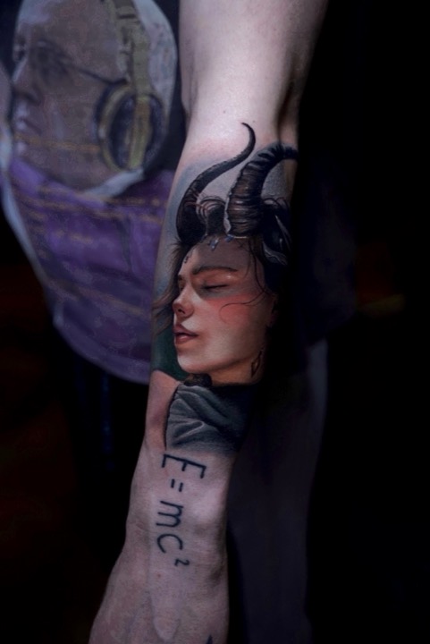 Woman with curled black horns and closed eyes, E=mc² written below, on a forearm.