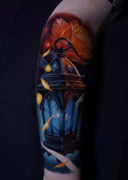 Blue glowing lantern with orange autumn leaves and drifting paper, colour tattoo on an arm.