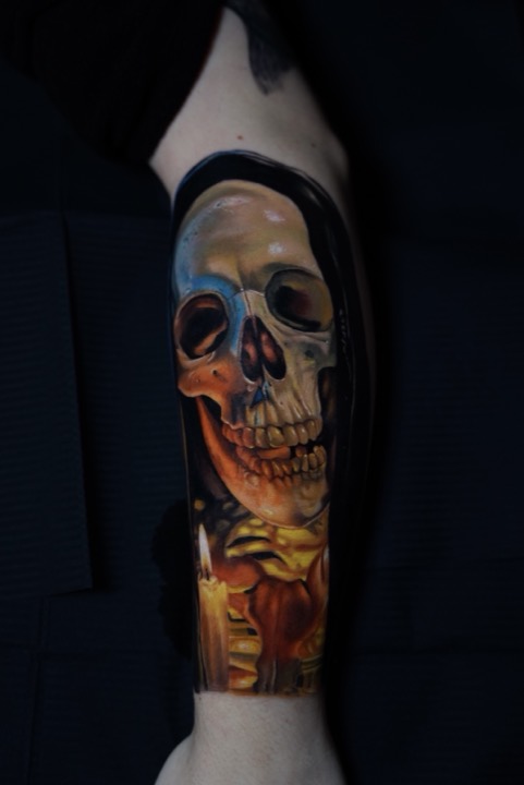 Skull lit by a candle in warm yellow and cold blue, colour tattoo on a forearm.