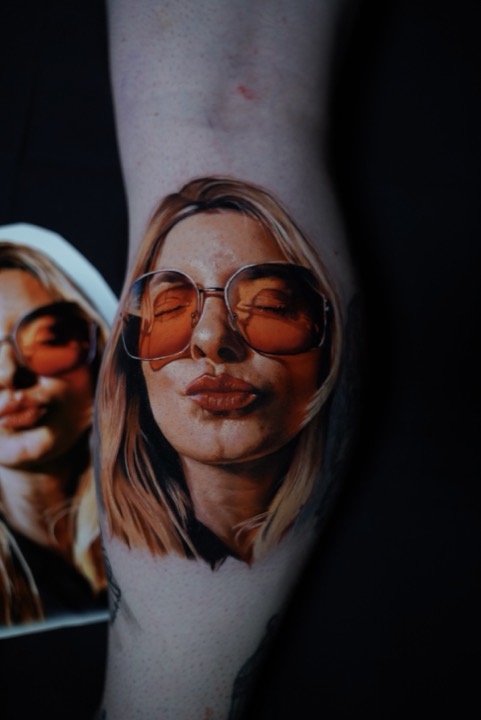 Woman in orange-tinted round sunglasses pouting, tattooed on a forearm next to the reference photo.