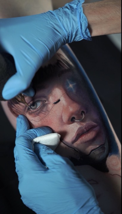 Blue-gloved hands tattooing and wiping a colour portrait of a woman, the lower edge still in outline.